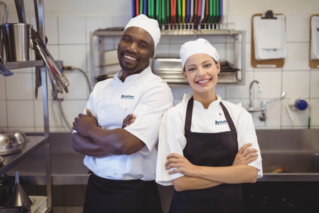 Man and woman with arms crossed smiling in kitchen, wearing HCSG Chef attire.