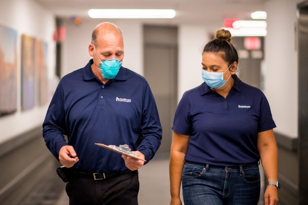 Two HCSG employees walking down a facility hallway, wearing masks and looking at a clipboard in HCSG employee attire.