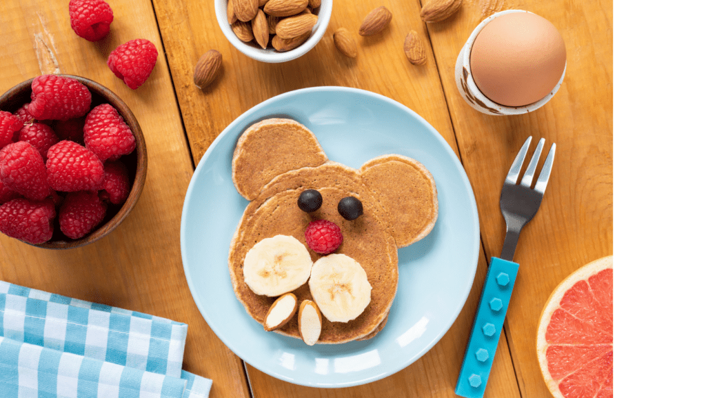 An aerial image of a breakfast meal assortment including a bowl of raspberries, almonds, and. a hard-boiled egg. The focal point is a pancake with a cartoon bear's face one it made out of various fruit.