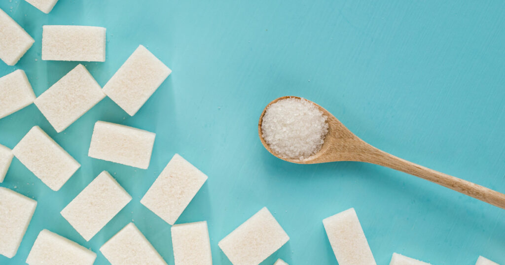A birds-eye photo of a wooden spoon filled with sugar next to a sugar cubes on a teal background.