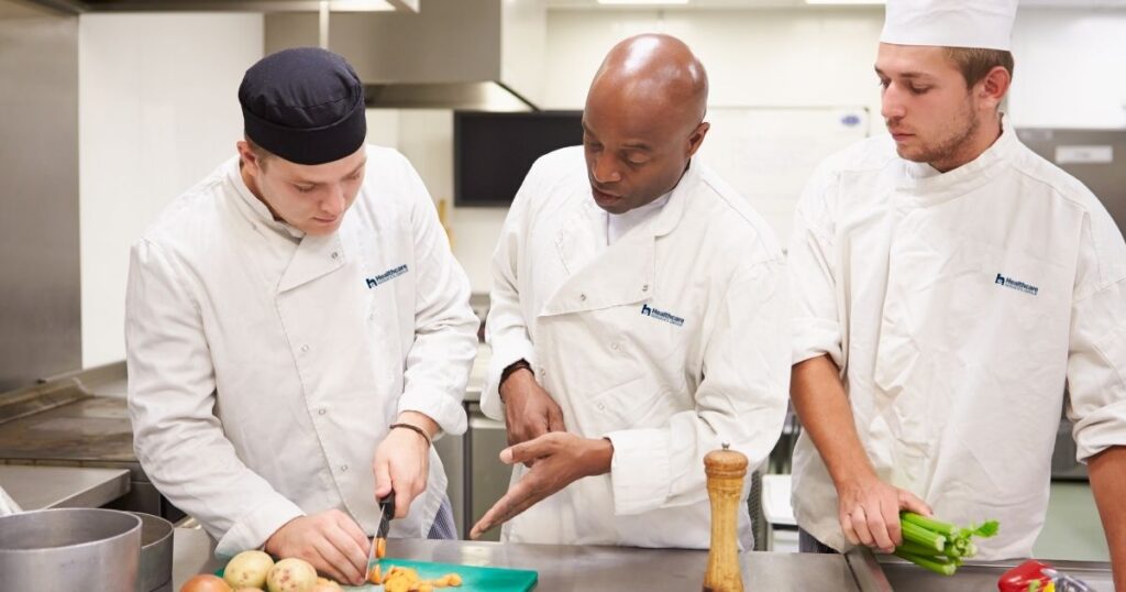 Three men in a facility kitchen wearing HCSG chef uniforms. One man is chopping vegetables, while another is directing him, and the third man looks upon the lesson.
