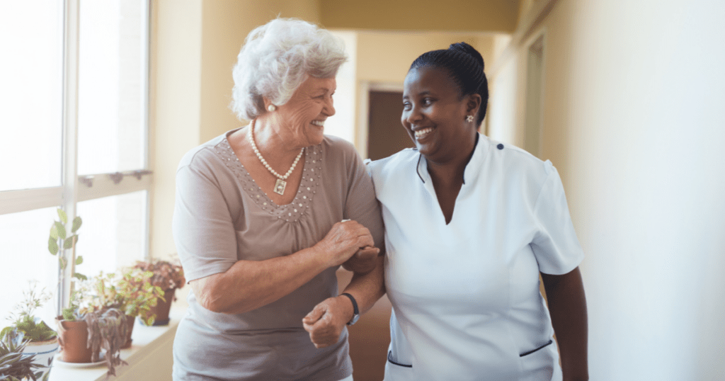 Nurse and resident laugh together, arm in arm, while walking down a sunlit facility hallway.