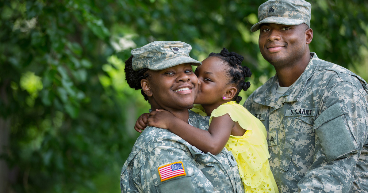 A man and woman smiling, dressed in US Army Uniforms. The woman is holding a a young girl who is kissing her cheek and smiling.