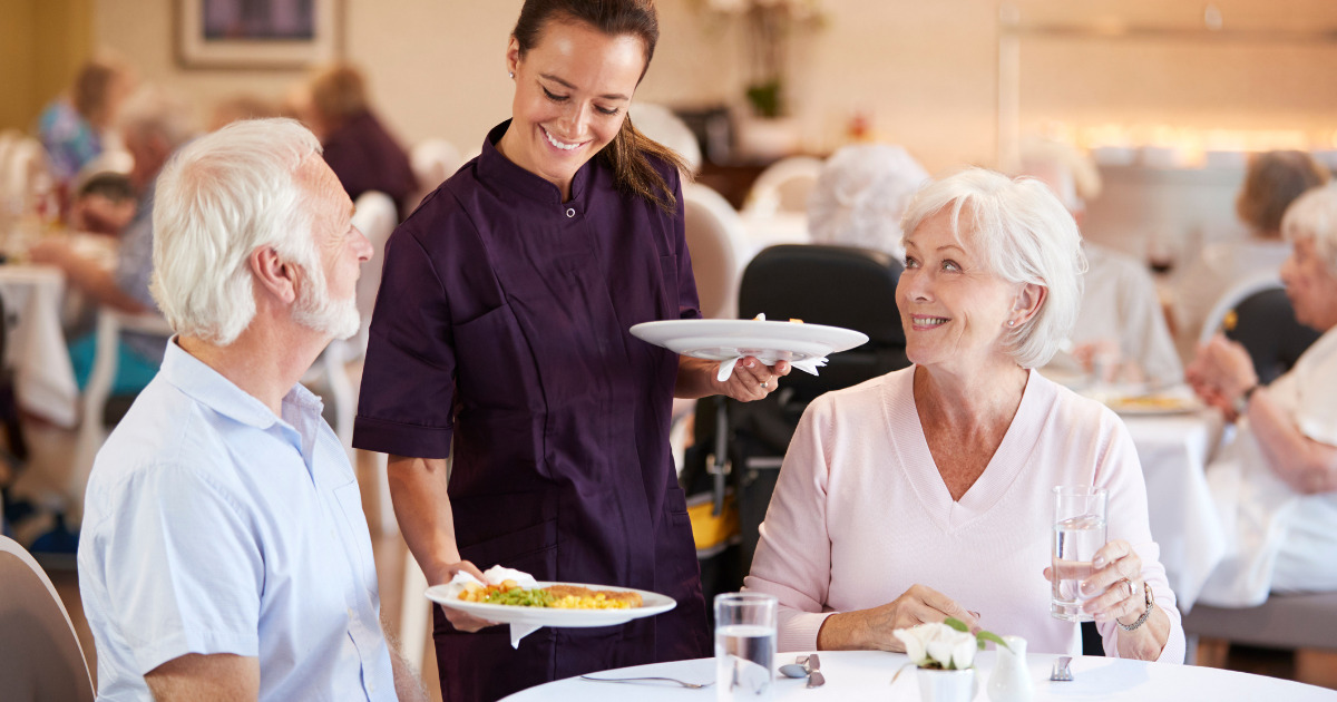 Dining services team member serving elderly man and woman in facility dining room.